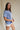 Upper body side view of female model wearing the Coastal Cowgirl Light Blue Graphic Tee that has light blue fabric, a white cowgirl boot graphic, white "Coastal Cowgirl" text, short sleeves, and a round neck.