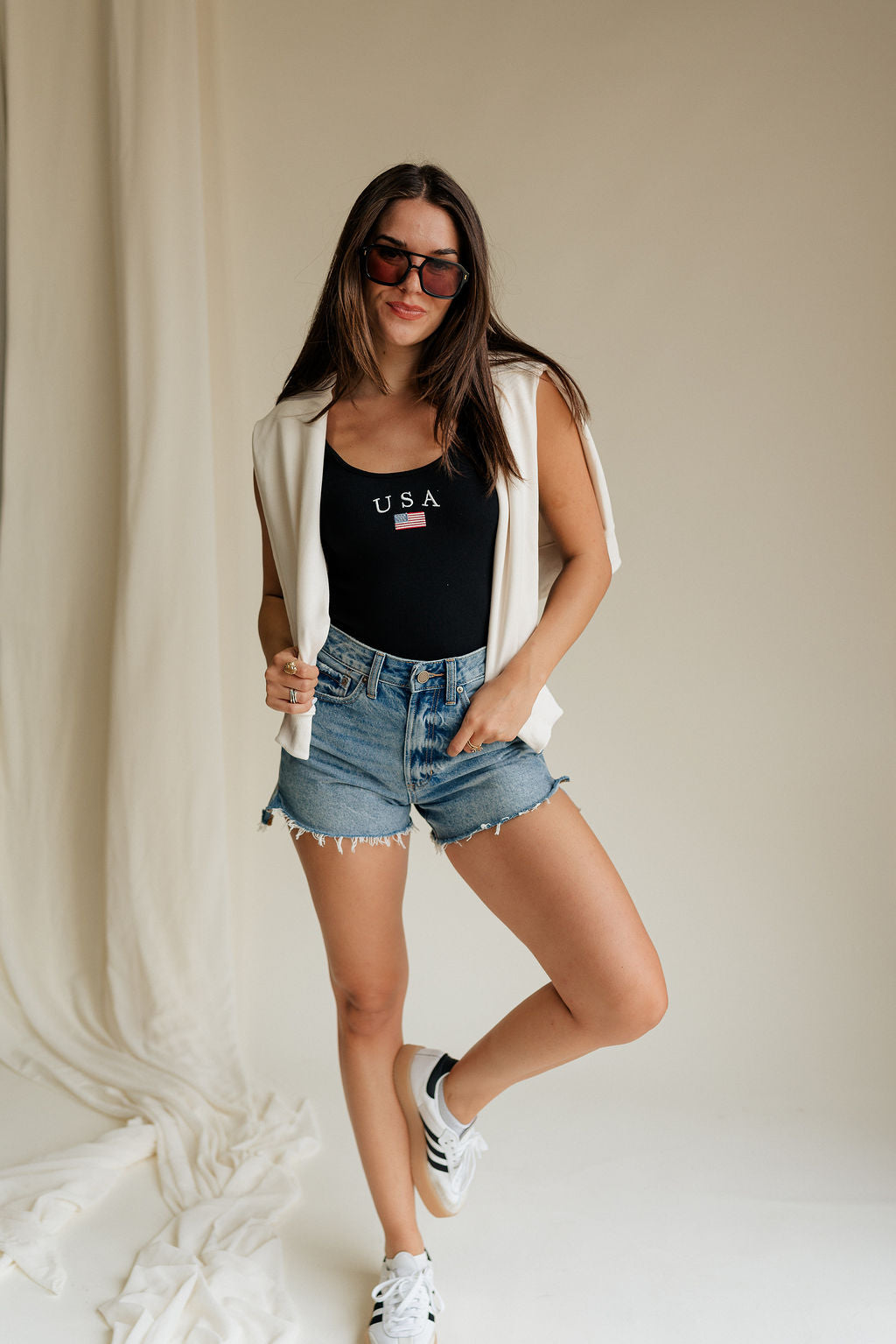Full body front view of female model wearing the USA Black Ribbed Sleeveless Bodysuit that has black ribbed fabric, USA in white text above a flag graphic, and a scoop neck. Styled with ivory sweatshirt around shoulders and denim shorts.