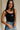 Upper body front view of female model wearing the USA Black Ribbed Sleeveless Bodysuit that has black ribbed fabric, USA in white text above a flag graphic, and a scoop neck. Styled with ivory sweatshirt around shoulders and denim shorts.