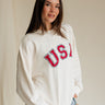 Upper body side view of female model wearing the USA Ivory Graphic Sweatshirt that has ivory fabric, USA written in red letters, long sleeves, and a round neck.