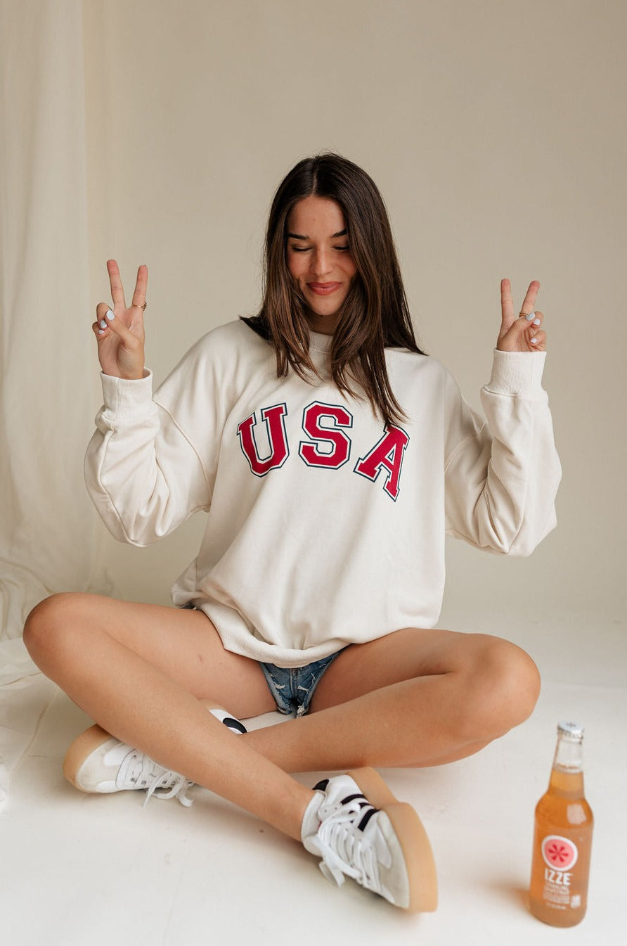 Upper body front view of female model wearing the USA Ivory Graphic Sweatshirt that has ivory fabric, USA written in red letters, long sleeves, and a round neck. Model is doing peace signs and sitting.