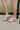 Side view of model's legs; model is wearing the Dolce Vita Notice Sneaker in White/Grey Leather that have white leather uppers with grey details, and a brown rubber sole.