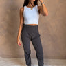 Full body front view of female model wearing the Uma Charcoal Jogger Pants that have charcoal grey fabric, a wide elastic waistband, tapered ankles, and side & back pockets.