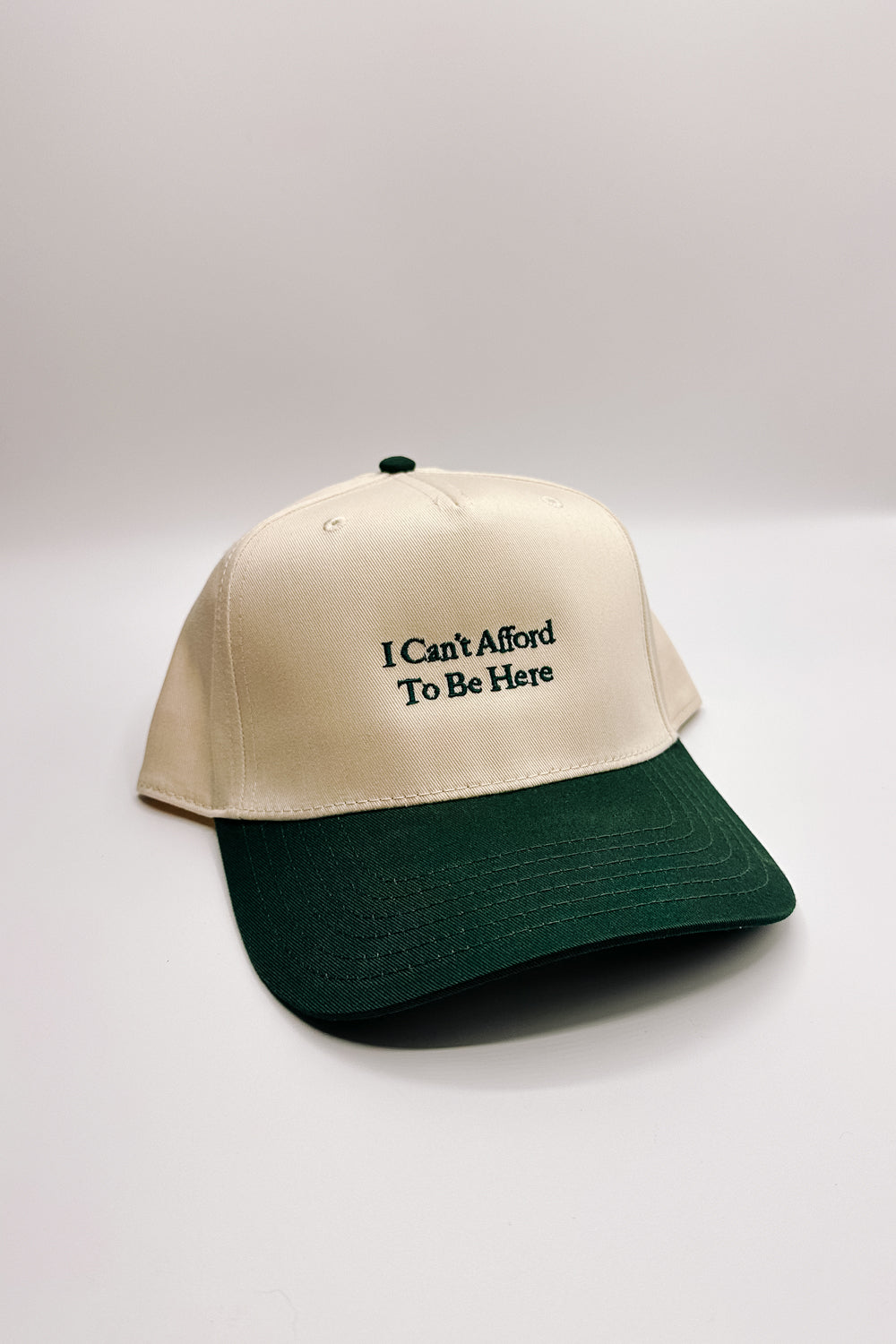 Image shows front view of the I Can't Afford To Be Here Trucker Hat against a white background.