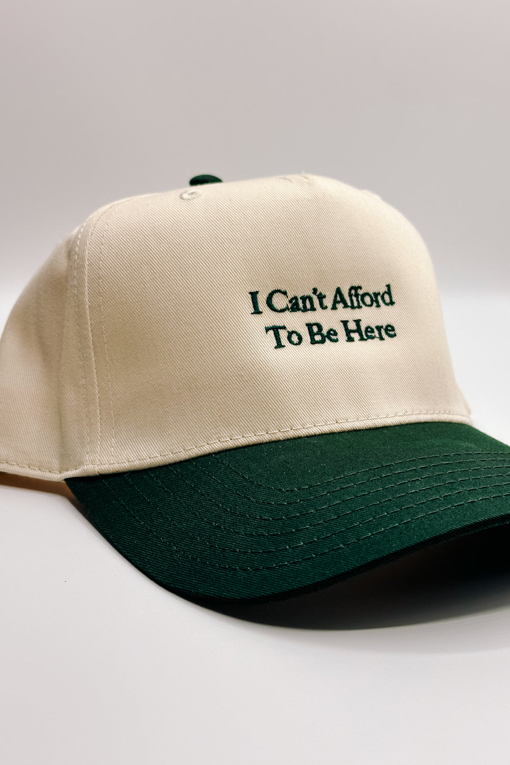 Image shows front view of the I Can't Afford To Be Here Trucker Hat against a white background. 