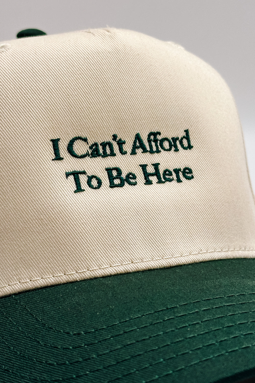 Image shows close front view of the I Can't Afford To Be Here Trucker Hat against a white background.