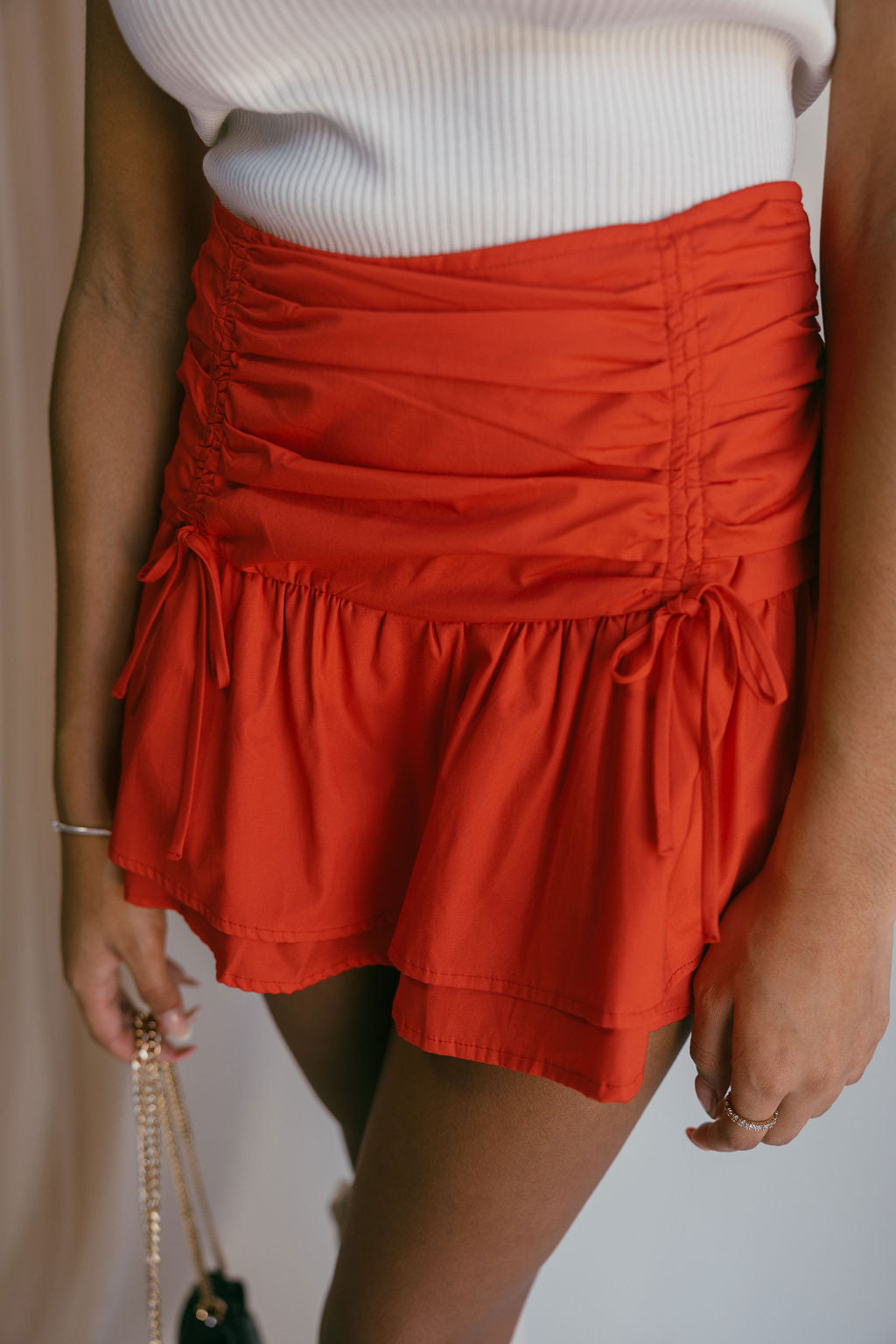 Close up front view of female model wearing the Calista Red Ruched Mini Skirt that has red fabric, ruched sides, and tiers. Worn with white tank top.