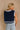 Close up back view of model wearing the Amanda Navy Cropped Sleeveless Sweater. This is a navy knit sweater, cropped, sleeveless, white trimming, bows on sides.