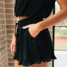 Close side view of female model wearing the Madeline Black Plisse Shorts that have black plisse fabric, elastic waist, pockets, and front tie. Worn with matching top