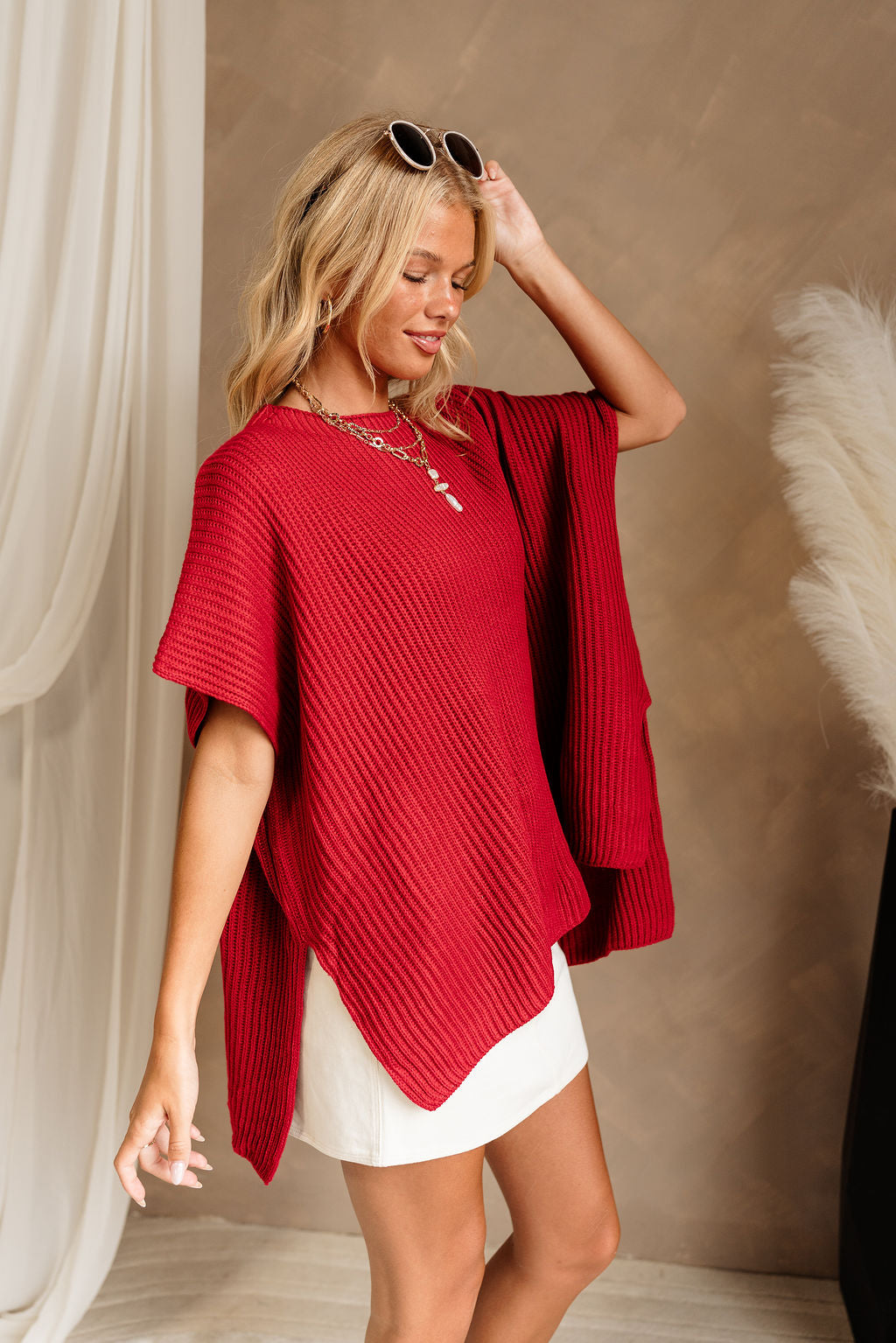 Side view of female model wearing the Heather Oversized Sweater in Red, which has a round neck, side slits, and short poncho sleeves. 