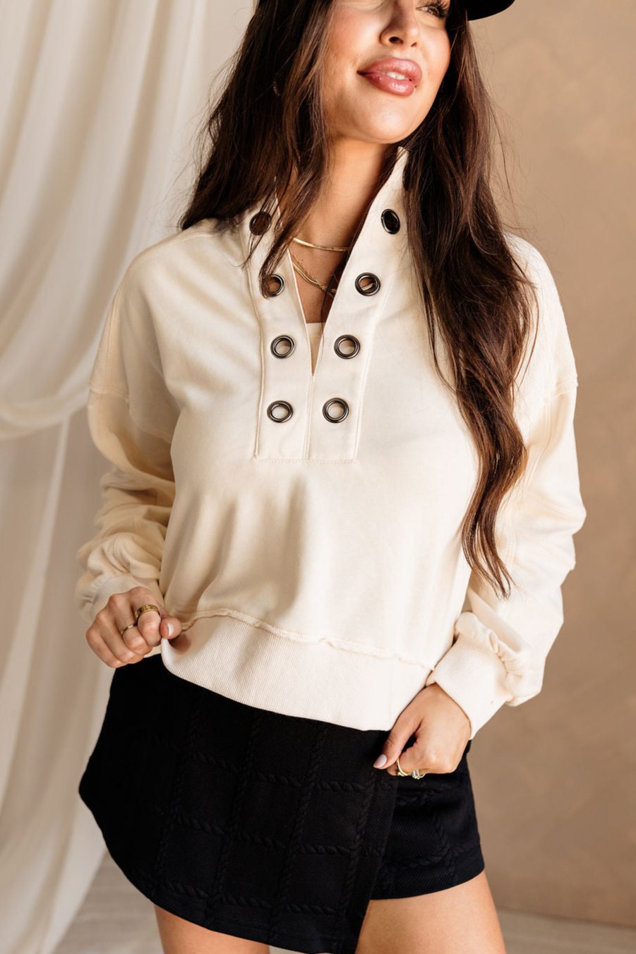 Front view of female model wearing the Caitlin Cream Pullover that has mixed cream fabrics, a grommet neckline, and long sleeves. Worn over black skort.