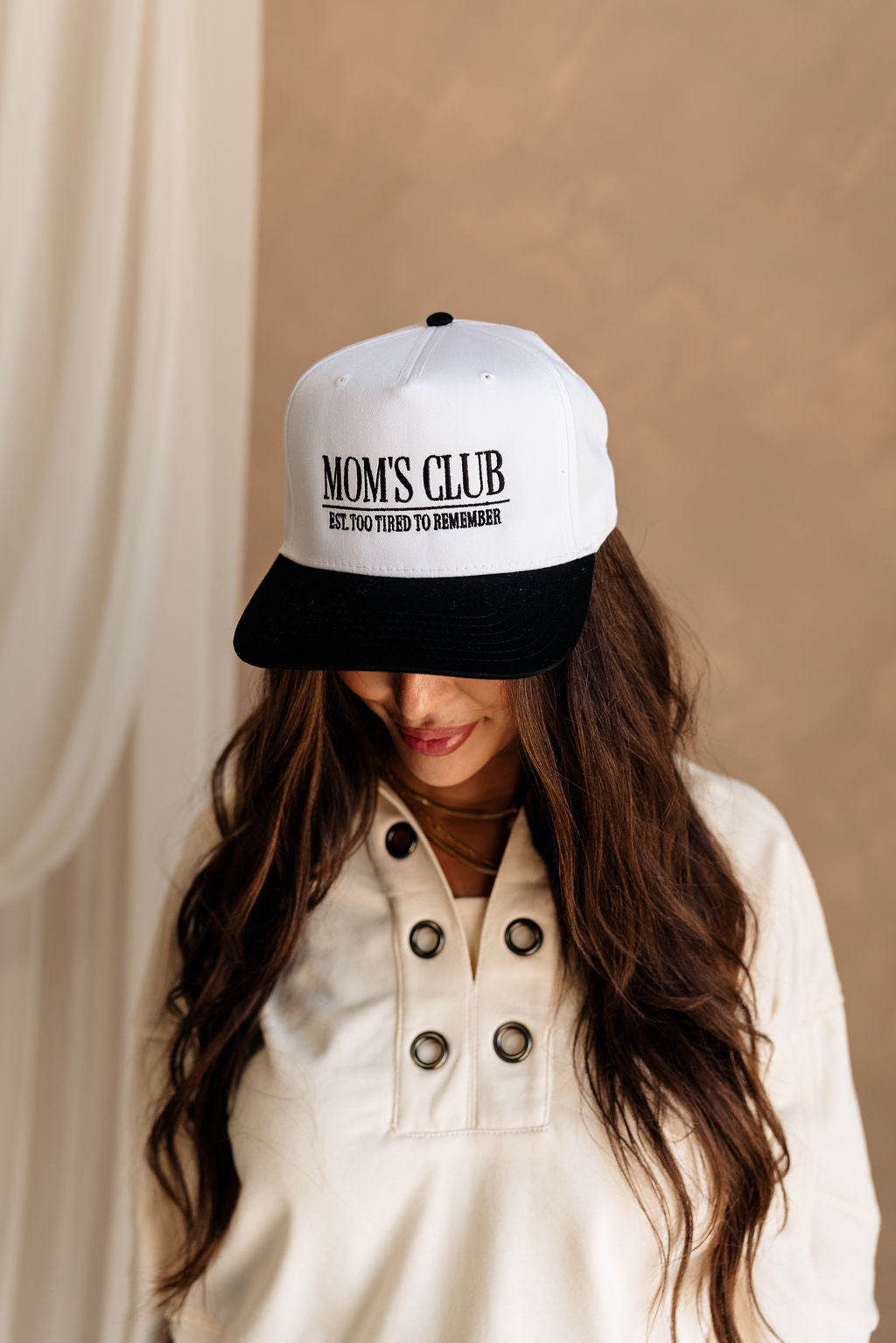 Front view of female model wearing the Mom's Club Trucker that is white with a black bill and text. Text on front says "MOM'S CLUB EST. TOO TIRED TO REMEMBER" and the back says "The happiest :)"