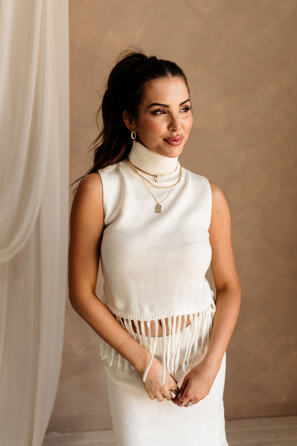 Upper body front view of female model wearing the Hillary Ivory Fringe Sweater Tank that has ivory knit fabric, a turtleneck, and a fringe hem. Worn with matching skirt.