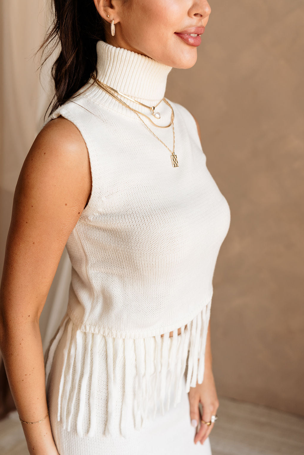 Upper body side view of female model wearing the Hillary Ivory Fringe Sweater Tank that has ivory knit fabric, a turtleneck, and a fringe hem. Worn with matching skirt.