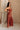 Full body side view of female model wearing the Carmella Rust Wide Leg Jumpsuit that has rust fabric, crochet trim, and wide legs.