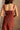 Upper body back view of female model wearing the Carmella Rust Wide Leg Jumpsuit that has rust fabric, crochet trim, and wide legs.