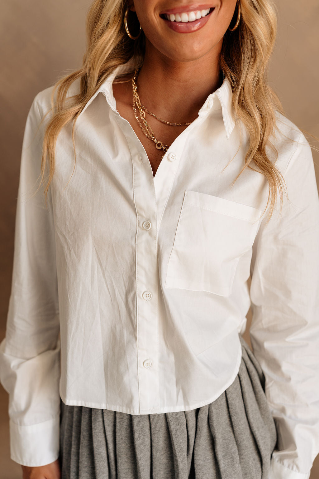 Close up front view of female model wearing the Claudia White Cropped Button Up that has a cropped waist, long sleeves, and button front with collar. Worn with grey skirt.