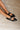 Image shows model's leg and feet against a cream rug. Model is wearing the Livia Black Patent Buckle Flats that have black patent material, pointed toles, and silver buckle details.