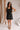 Full body front view of female model wearing the Peyton Denim Zip Front Mini Dress in black, that has washed black denim fabric, a gold front zipper, straps with buckle adjustments, and pockets.
