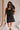 Front view of female model wearing the Peyton Denim Zip Front Mini Dress in black, that has washed black denim fabric, a gold front zipper, straps with buckle adjustments, and pockets.