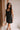 Frontal side view of female model wearing the Peyton Denim Zip Front Mini Dress in black, that has washed black denim fabric, a gold front zipper, straps with buckle adjustments, and pockets.