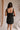 Back view of female model wearing the Peyton Denim Zip Front Mini Dress in black, that has washed black denim fabric, a gold front zipper, straps with buckle adjustments, and pockets.