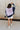 Full body back view of female model wearing the Lucia Color Block Sweatshirt that has purple, navy, and grey color blocking. Worn with white shorts.