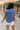 Upper body back view of female model wearing the Leah Blue Short Sleeve T-Shirt, paired with white shorts.