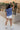 Full body back view of female model wearing the Leah Blue Short Sleeve T-Shirt, paired with white shorts.