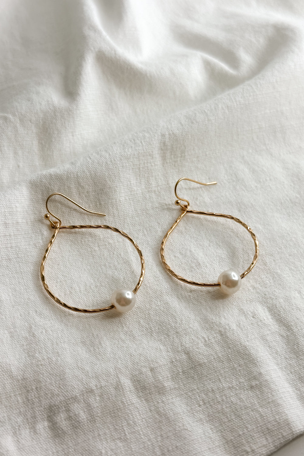 Flat lay view of the Elizabeth Gold Pearl Hoop Dangle Earring which features open hammered, gold hoop with single pearl detail