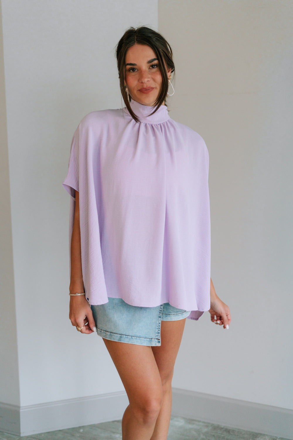 Front view of female model wearing the Madilyn Lavender High Neck Top which features Lavender Lightweight Fabric, High Neckline with Tie Closure and Short Sleeves