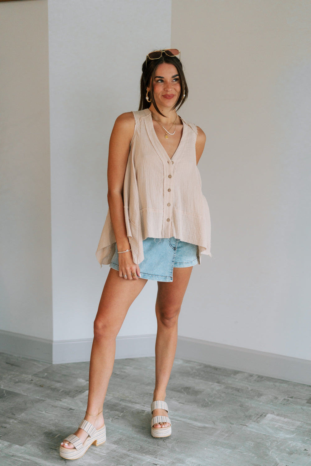 Full body view of female model wearing the Heidi Light Beige Gauze Sleeveless Top which features Taupe Gauze Fabric, Raw Hem, Wooden Button Up and V-Neckline