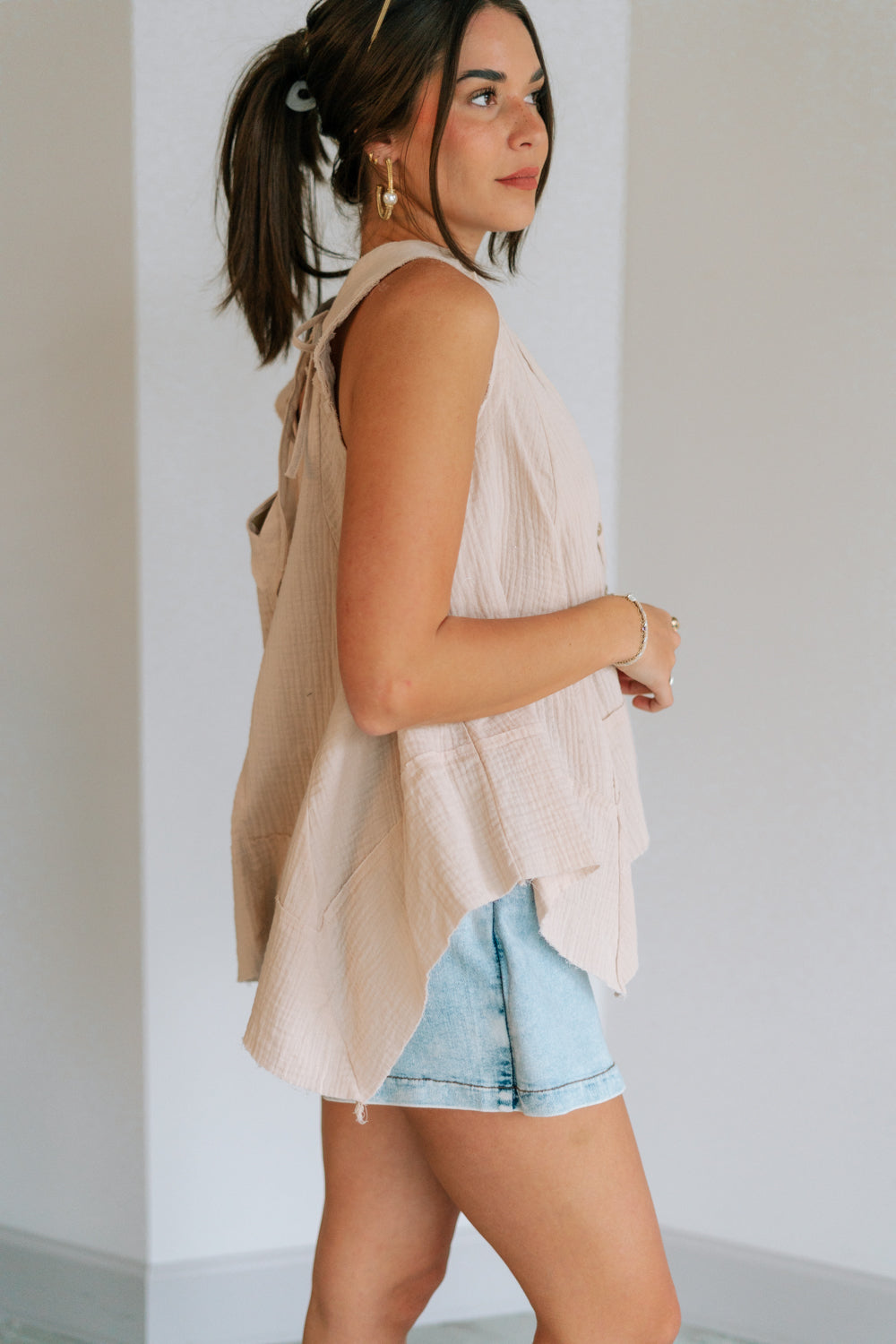 Side view of female model wearing the Heidi Light Beige Gauze Sleeveless Top which features Taupe Gauze Fabric, Raw Hem, Wooden Button Up and V-Neckline