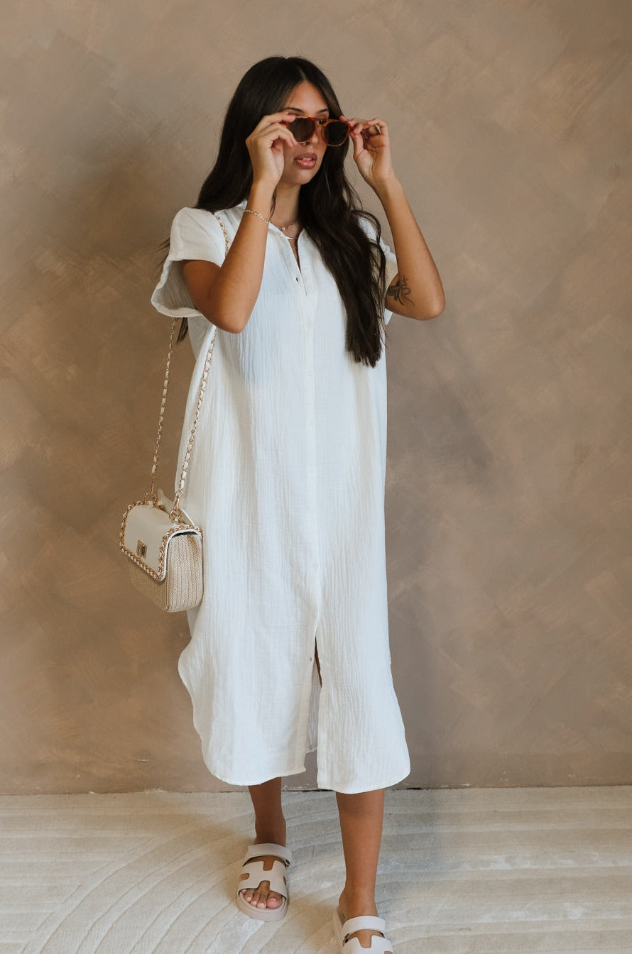 Full body front view of female model wearing the Maris White Button-Up Midi Dress that has white gauze fabric, button up front, short sleeves, and a collar. Worn white white sandals and  beige purse. Model is holding sunglasses