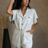 Front view of female model wearing the Lydia Ivory & Black Striped Romper that has white fabric with thin black stripes, pockets, buttons, a collar, and short sleeves. 