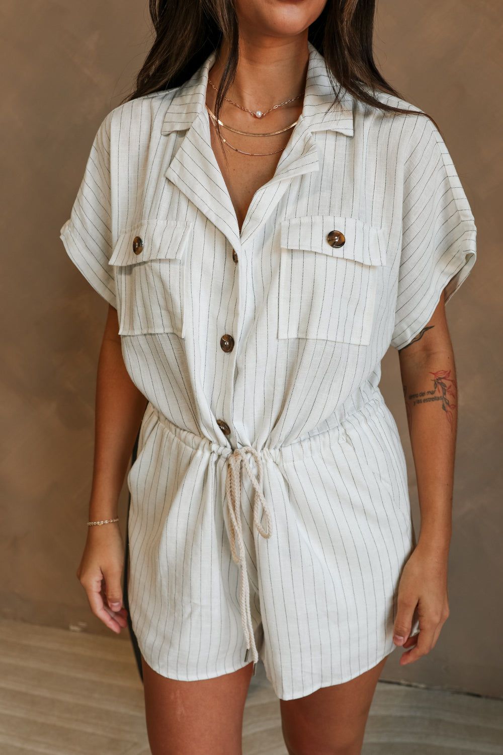 Close-up Front view of female model wearing the Lydia Ivory & Black Striped Romper that has white fabric with thin black stripes, pockets, buttons, a collar, and short sleeves. 