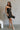 Full body front view of model wearing the Sarah Black Strapless Mini Dress that has black fabric with metallic black florals and a strapless neckline. Model is holding black purse; paired with cream platform heels.
