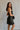 Side view of model wearing the Sarah Black Strapless Mini Dress that has black fabric with metallic black florals and a strapless neckline. Model is holding black purse