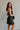 Side view of model wearing the Sarah Black Strapless Mini Dress that has black fabric with metallic black florals and a strapless neckline. Model is holding black purse