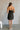 Full body back view of model wearing the Sarah Black Strapless Mini Dress that has black fabric with metallic black florals and a strapless neckline. paired with cream platform heels.