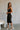 Full body side view of model wearing the Gianna Black & White Midi Dress that has black fabric, and white straps with white neckline bow trim. Model is wearing white heels