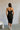Full body back view of model wearing the Gianna Black & White Midi Dress that has black fabric, and white straps with white neckline bow trim. Model is wearing white heels 