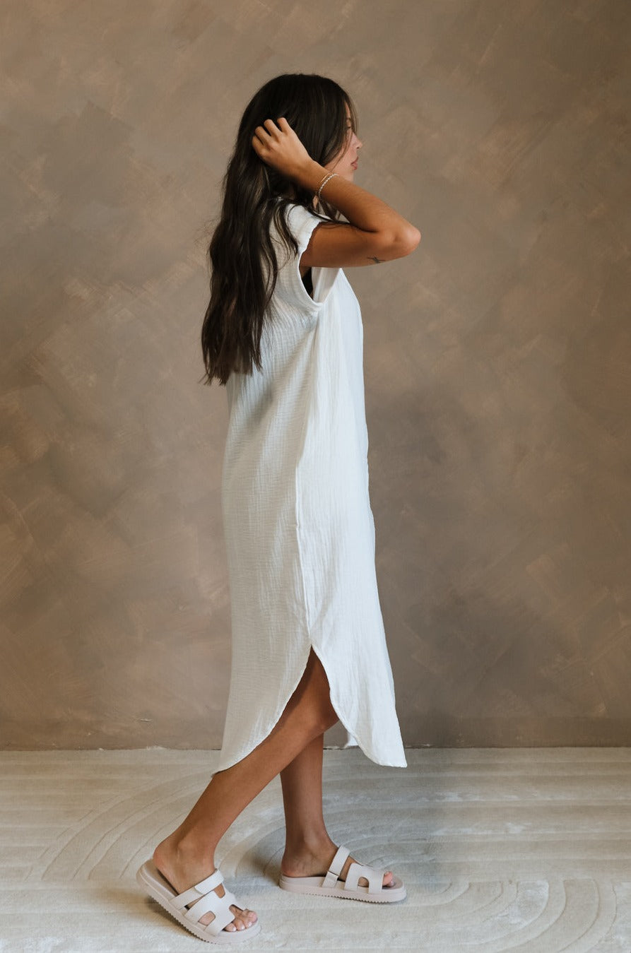 Full body side view of female model wearing the Maris White Button-Up Midi Dress that has white gauze fabric, button up front, short sleeves, and a collar. Worn white white sandals.