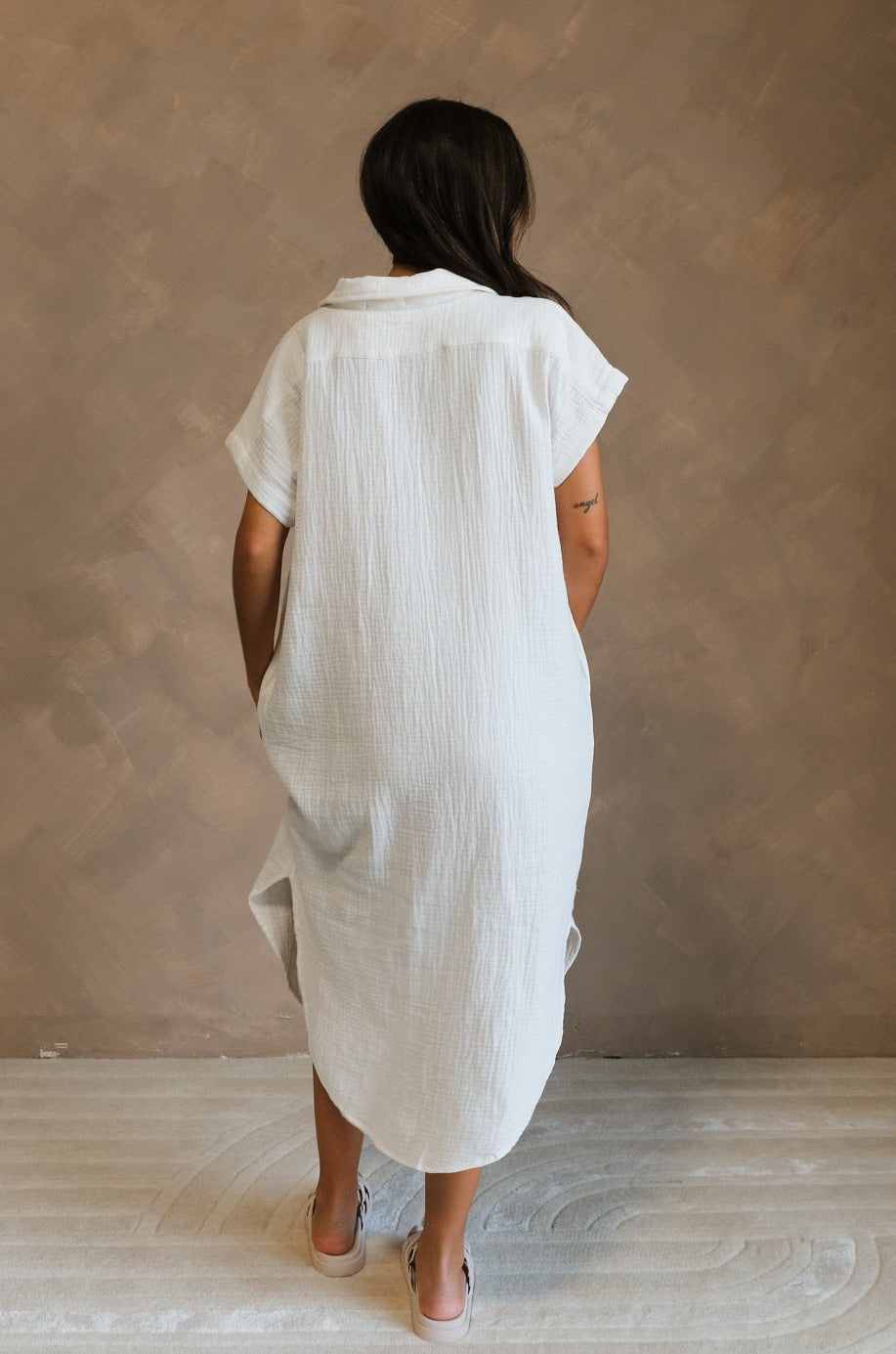 Full body back view of female model wearing the Maris White Button-Up Midi Dress that has white gauze fabric, button up front, short sleeves, and a collar. Worn white white sandals.