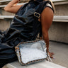 Side view of female model with the Johanna Clear Leopard Purse on her shoulder. Model is sitting on bleachers and wearing a black dress