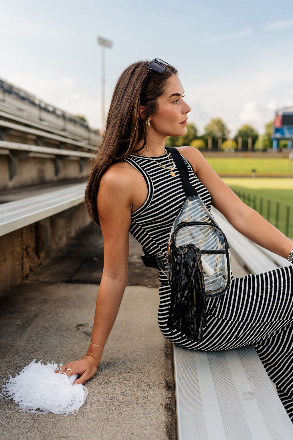 Image shows side view of model wearing the Riley Clear & Black Sling Bag. Model is sitting on bleachers and wearing a striped outfit.