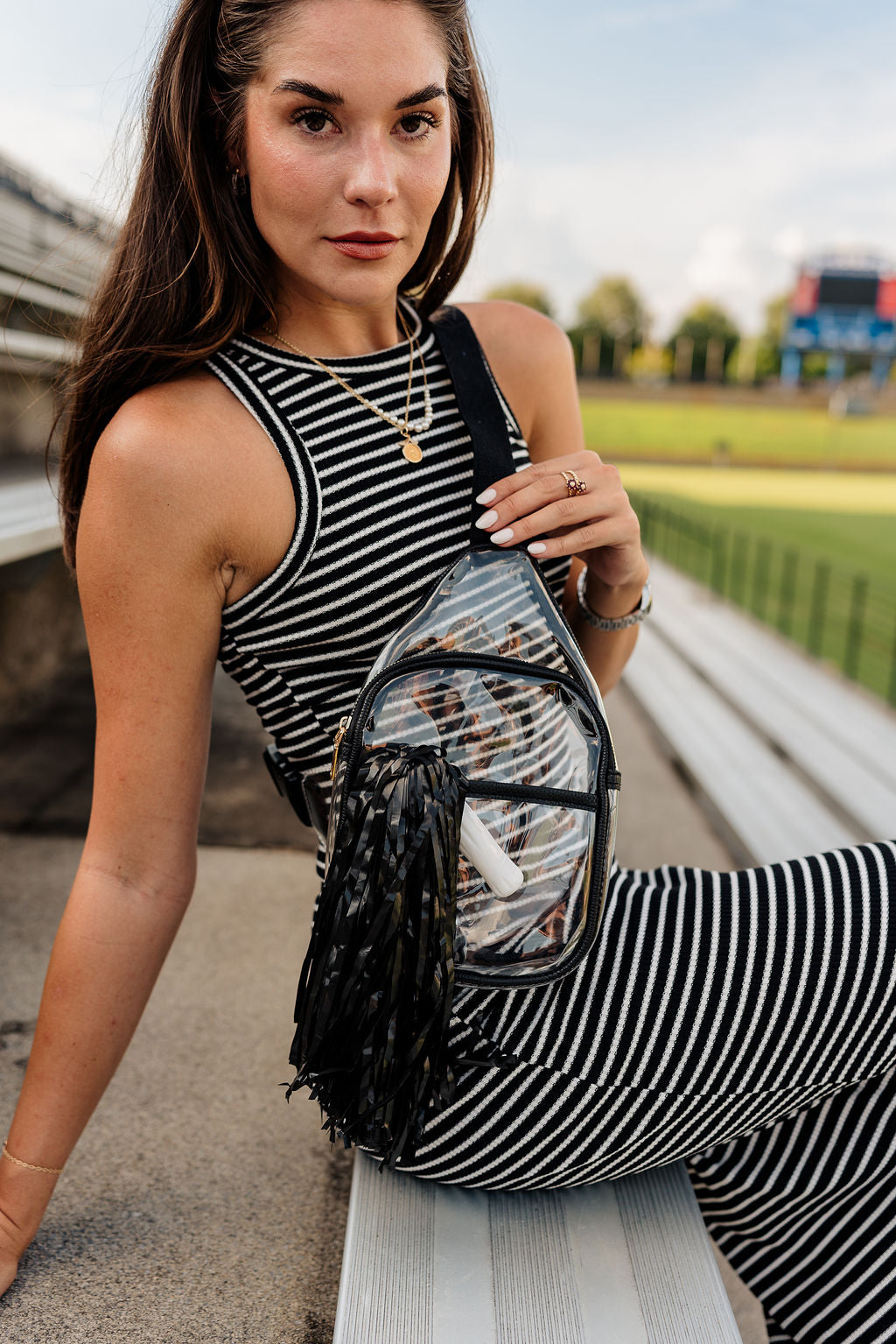 Image shows side view of model wearing the Riley Clear & Black Sling Bag. Model is sitting on bleachers and wearing a striped outfit.