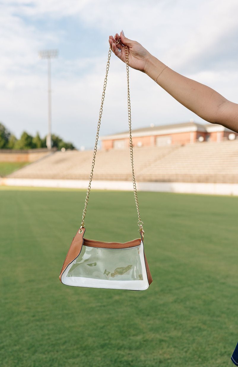 Model's hand is holding the Rebecca Clear Crossbody Purse in Tan in front of a grass field background.