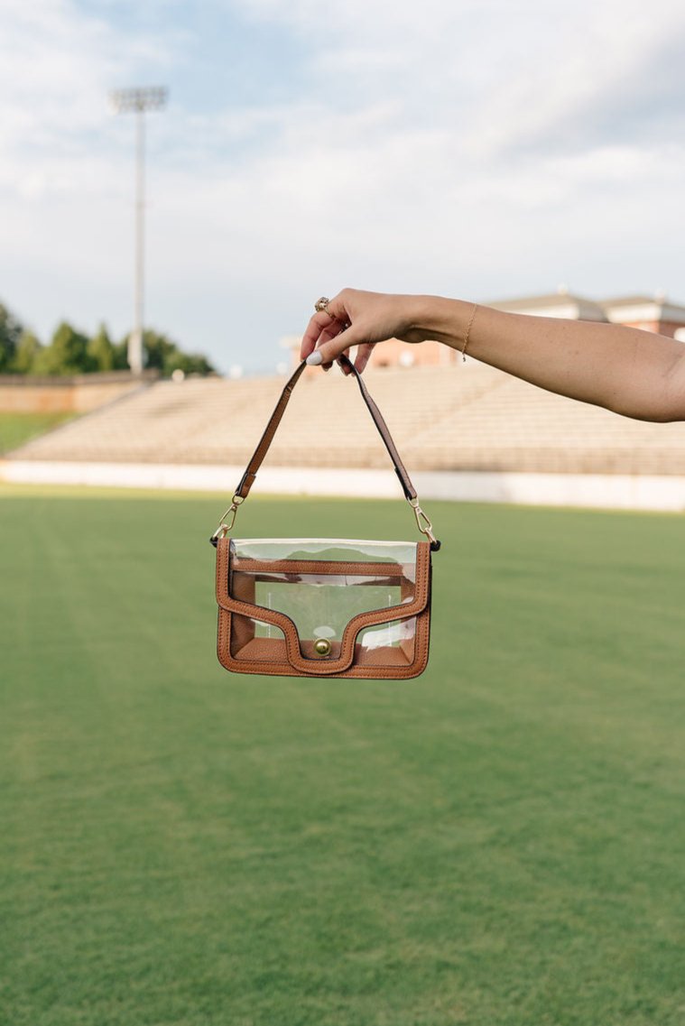 Model's hand is holding the Miley Clear Purse in Brown against a grass background.