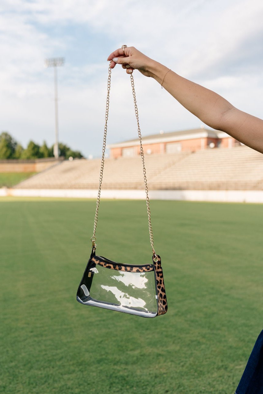 Model's hand is holding the Rebecca Clear Crossbody Purse in Leopard in front of a grass field background.